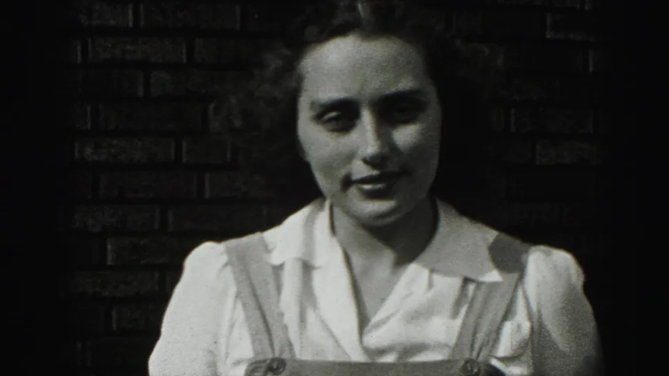 The woman appears in a similar close view, facing the camera with a slight smile against dark brickwork in the 1975 home movie.