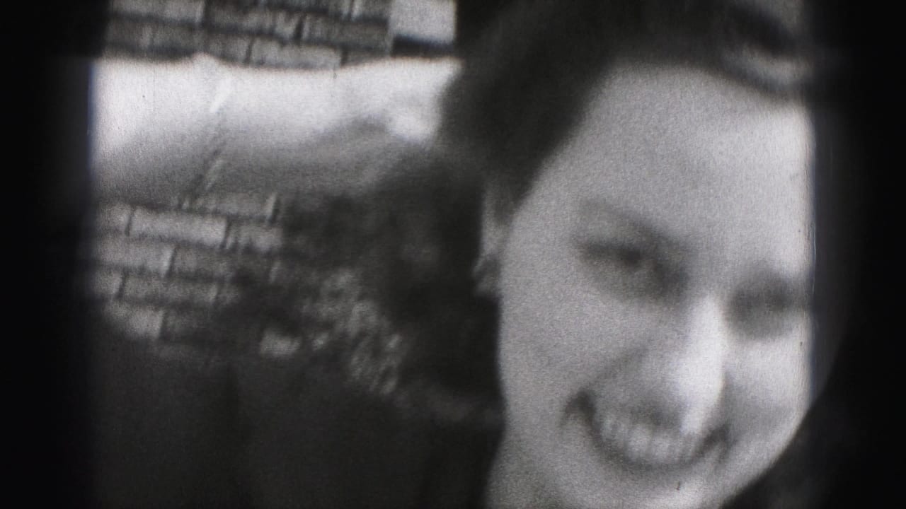 Young woman smiles toward the camera while raising an arm near a brick wall.