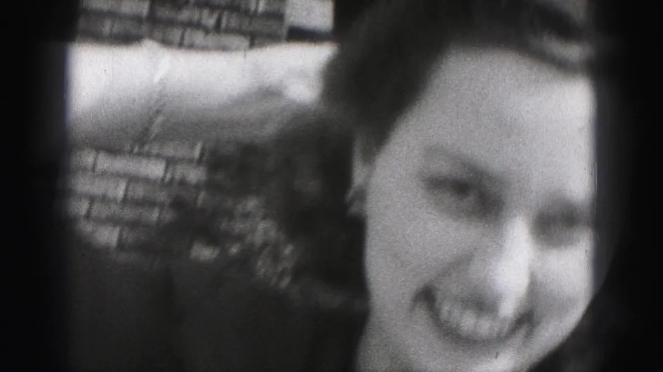 In this 1975 Harrisburg home-movie frame, a young woman smiles toward the camera with one arm lifted beside a brick wall.