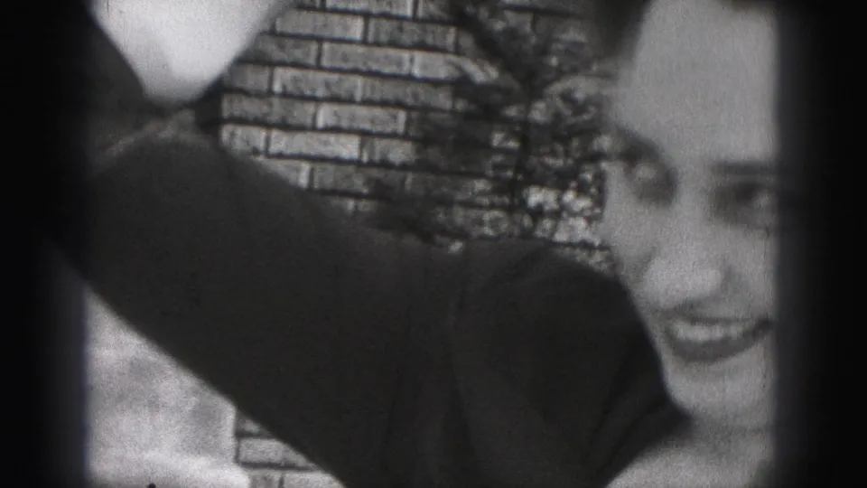The woman turns partly in profile and smiles, her arm raised as she adjusts her hair against the brick-wall background.