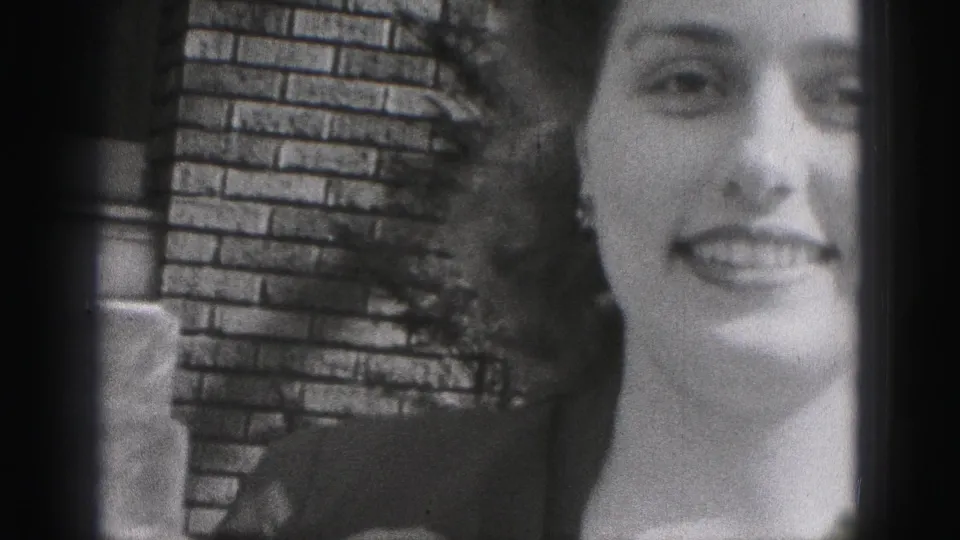The woman faces the camera in a soft black-and-white 8mm frame, with earrings and a brick wall visible behind her.