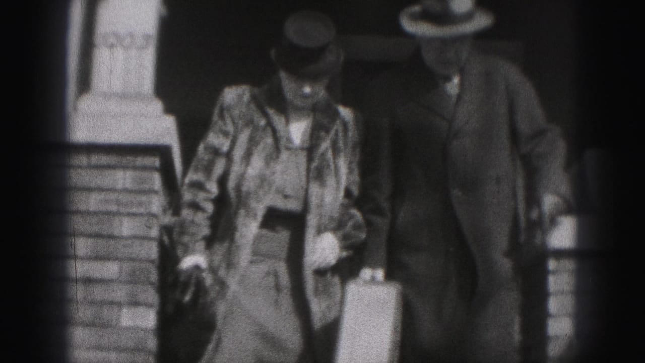 Two adults in hats walk beside a brick building, one carrying a suitcase.
