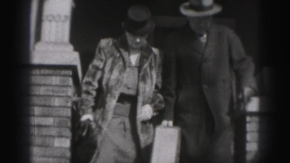 In 1975 Harrisburg home-movie footage, two adults in period dress walk outside a brick building with a suitcase.