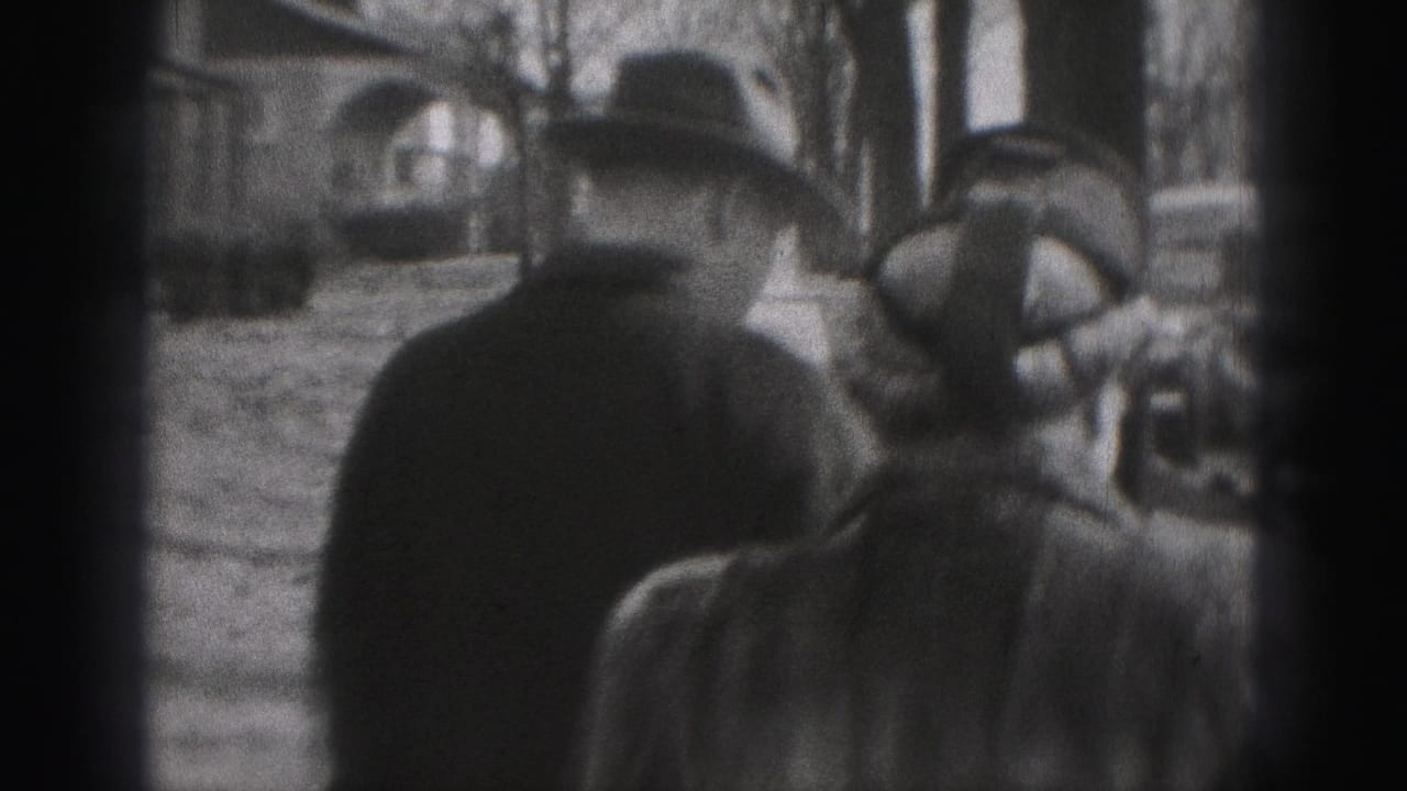 In supported 1975 Harrisburg home-movie footage, two adults in coats and hats walk away from the camera along a tree-lined sidewalk.