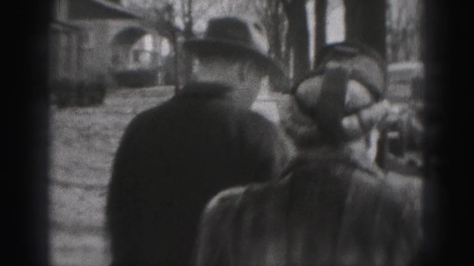 In supported 1975 Harrisburg home-movie footage, two adults in coats and hats walk away from the camera along a tree-lined sidewalk.