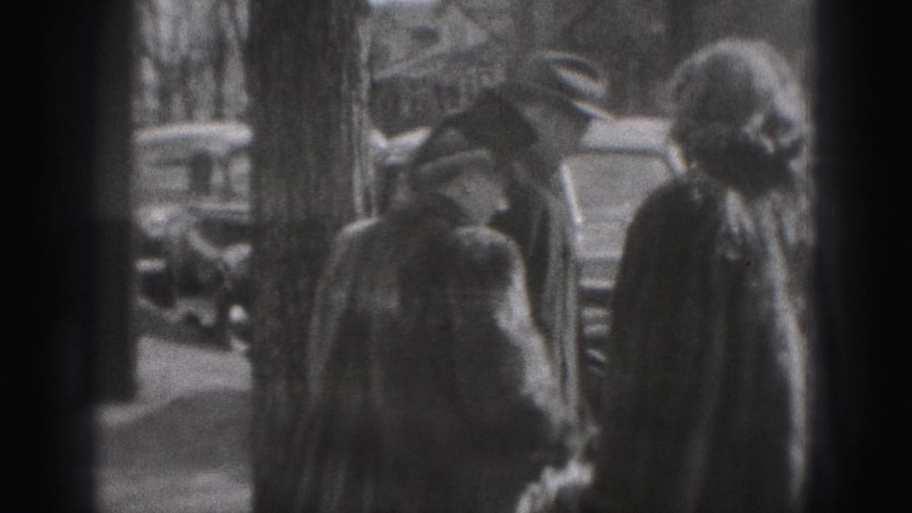 A woman in a coat and head covering stands near a man on a sidewalk, with cars and leafless trees visible behind them.