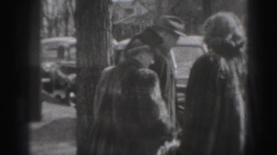 A woman in a coat and head covering stands near a man on a sidewalk, with cars and leafless trees visible behind them.