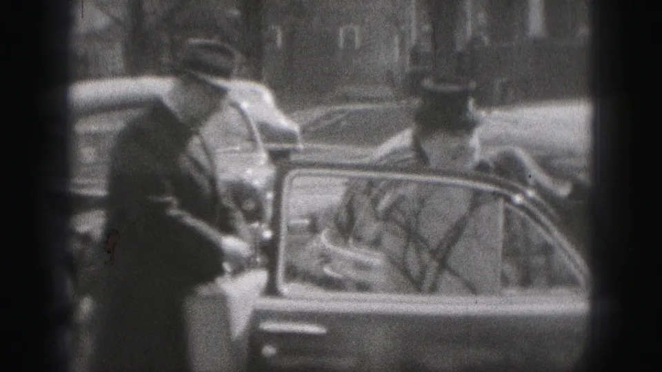 The man stands at an open car door as the couple reaches the vehicle, with reflections and bare branches visible in the window.