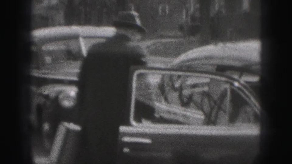 A man in a brimmed hat and dark coat stands next to the open car door, framed by grainy 8mm footage and street-side trees.