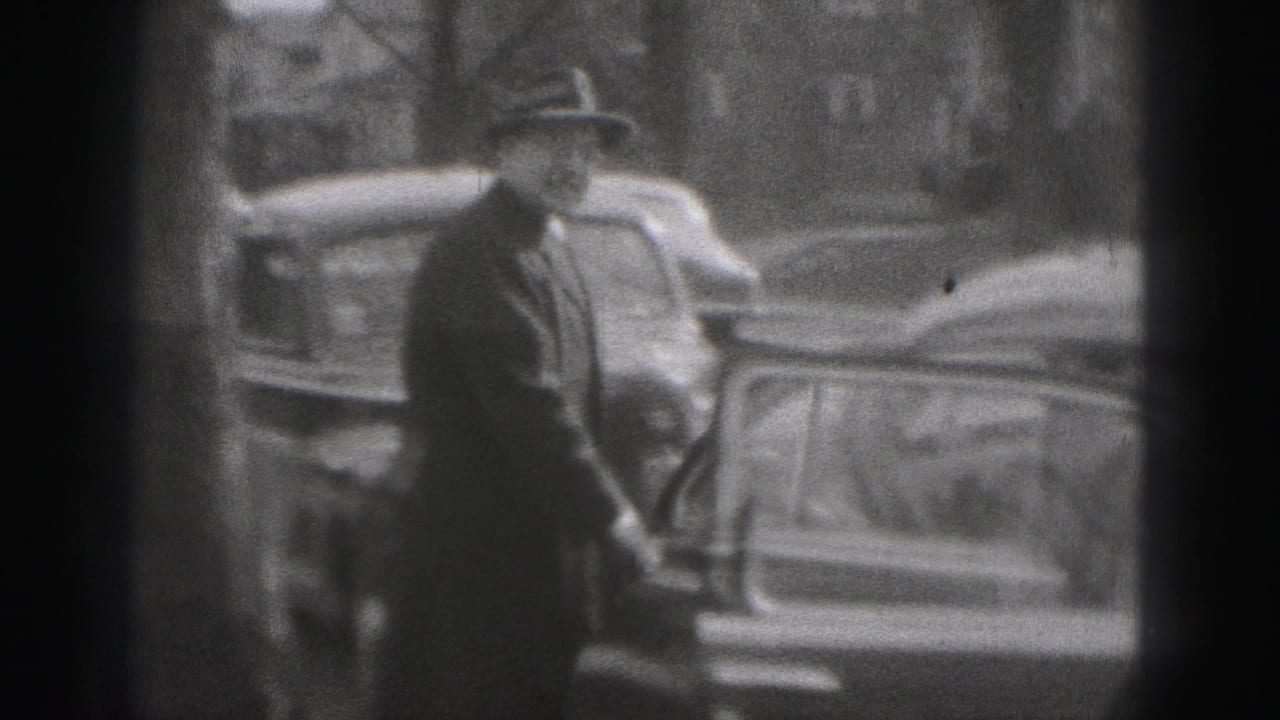 In Harrisburg 1975 footage, a man in a hat stands by a curbside car with other parked vehicles behind him.