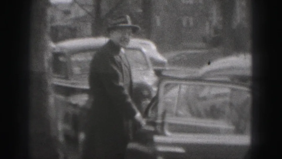In Harrisburg 1975 footage, a man in a hat stands by a curbside car with other parked vehicles behind him.