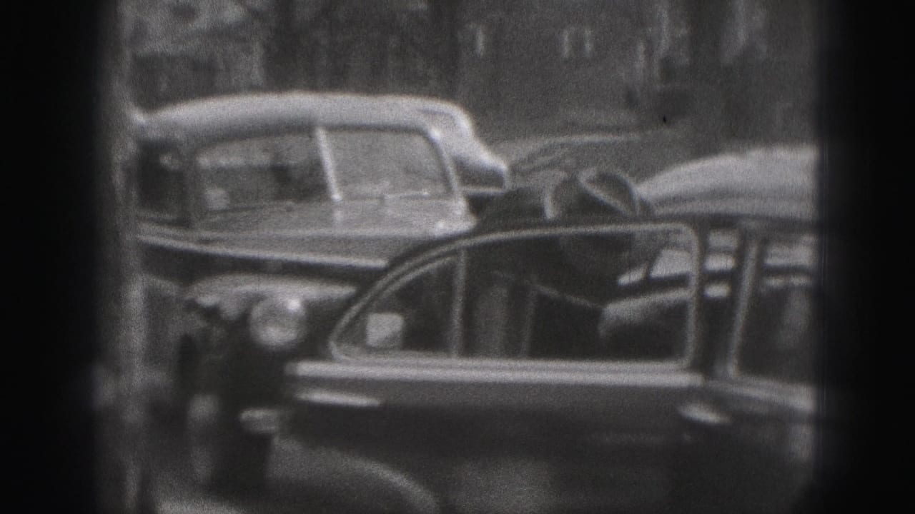 A car door stands open on a Harrisburg street, with parked sedans and blurred buildings in the 1975 home-movie frame.
