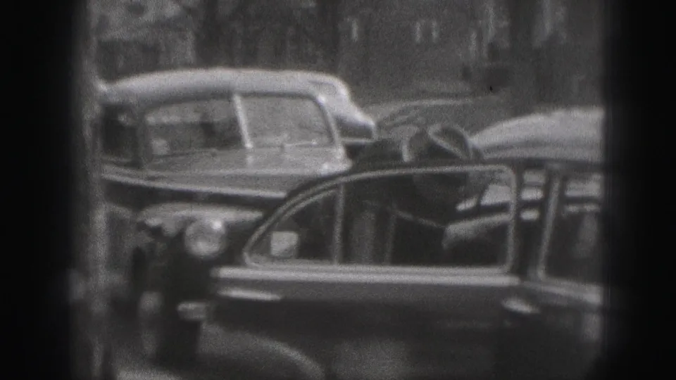 A car door stands open on a Harrisburg street, with parked sedans and blurred buildings in the 1975 home-movie frame.