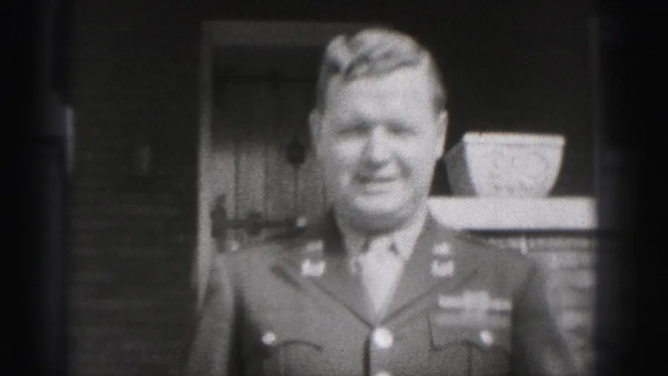 The man in dress uniform smiles close to the camera, with the front door, brickwork, and porch planter behind him.