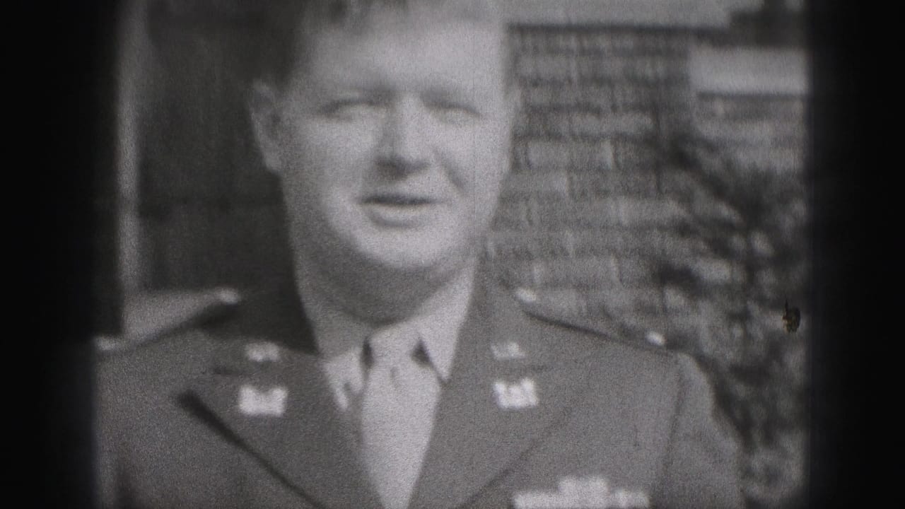 Adult man in a military-style uniform facing the camera outside a brick building.