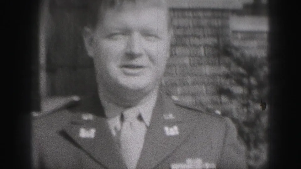 In a 1975 Harrisburg home-movie frame, an adult man in a military-style uniform faces the camera near a brick residential building.