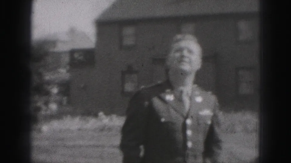 The uniformed man stands in a residential yard in Harrisburg, Pennsylvania, looking upward beside brick houses in soft 8mm footage.