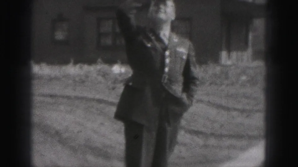 The man smiles while walking in uniform across a residential yard, with a brick house behind him in the 1975 Harrisburg footage.