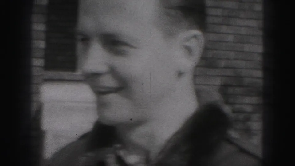 A smiling adult man appears in profile near a brick wall in 1975 Harrisburg home-movie footage, with soft focus and visible film grain.