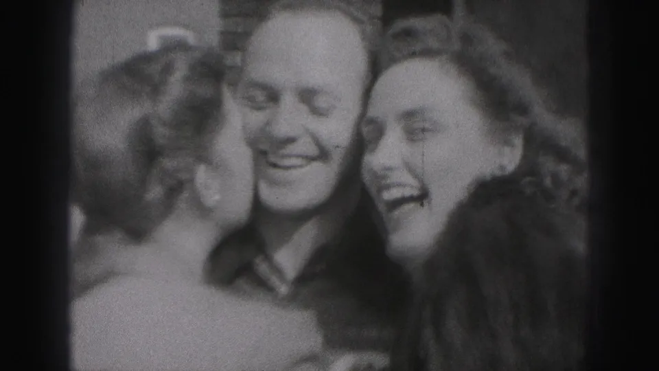 Two women lean toward a smiling man in a warm 1975 Harrisburg reunion moment captured on 8mm film.