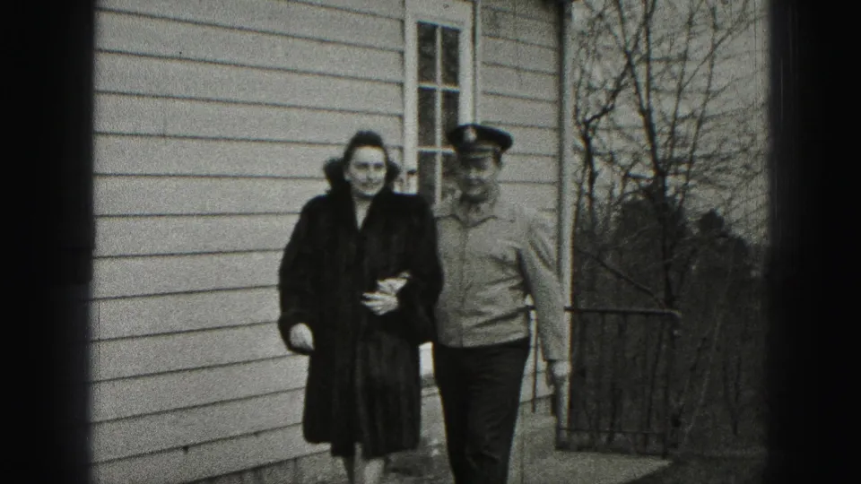 In Harrisburg, Pennsylvania home-movie footage, a woman in a dark fur coat walks beside a uniformed man along a residential walkway.