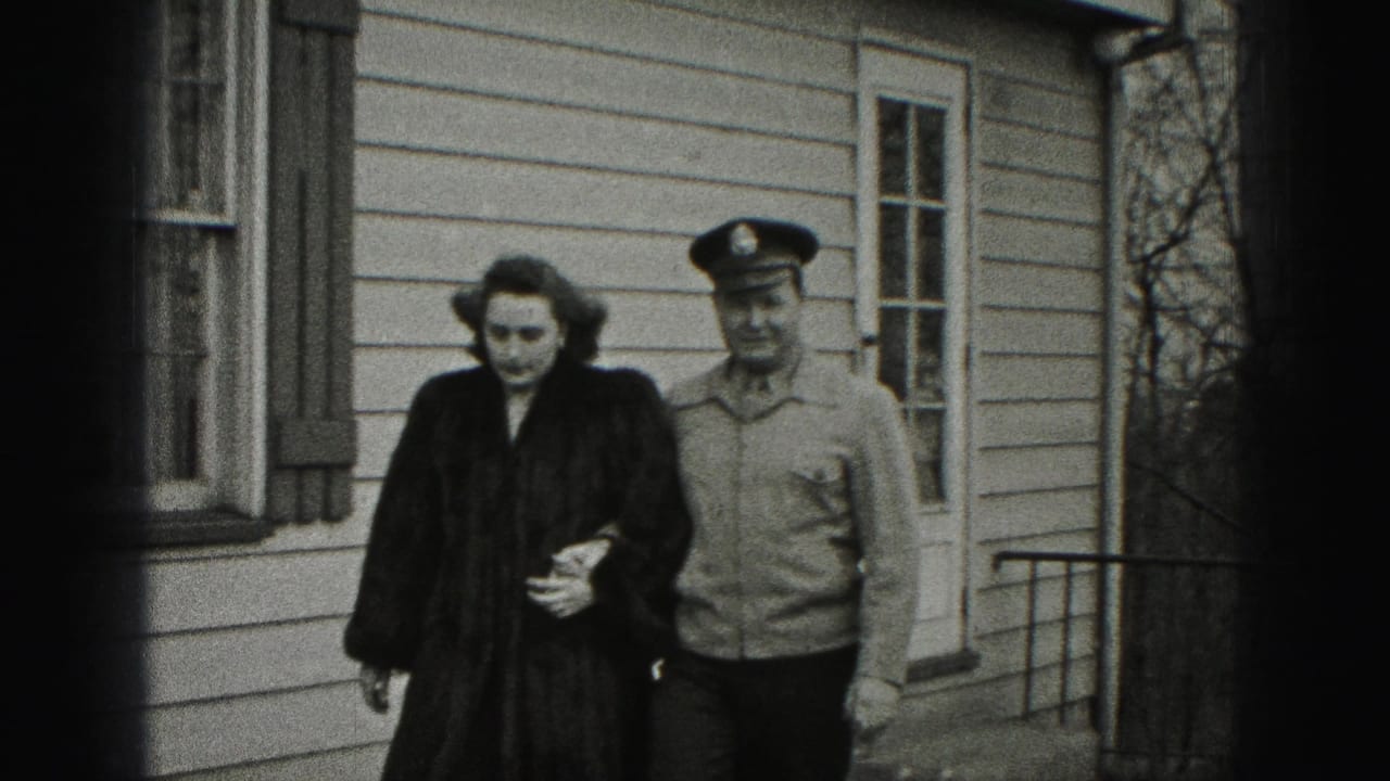 Uniformed man escorts a woman along a walkway in front of a house with windows and siding.