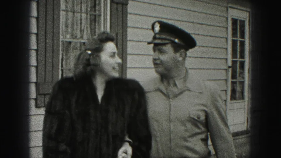 Near the end of the clip, the woman turns toward the uniformed man while they walk beside the residential exterior.