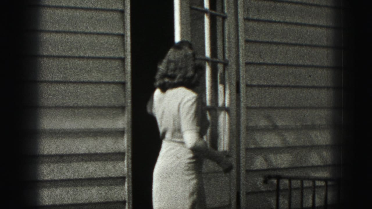 In Harrisburg, Pennsylvania home-movie footage, a woman in a light dress stands at an open exterior door beside horizontal siding.