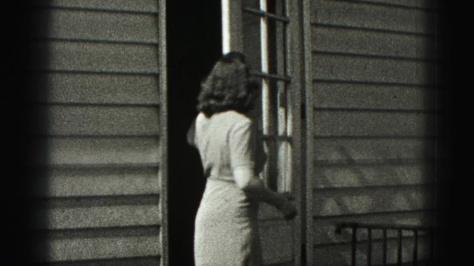 In Harrisburg, Pennsylvania home-movie footage, a woman in a light dress stands at an open exterior door beside horizontal siding.
