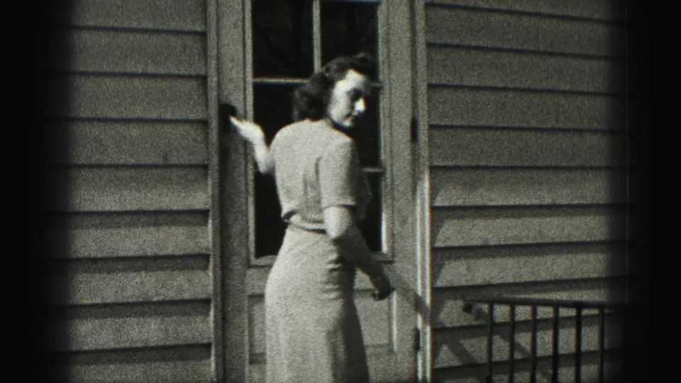 The woman turns back toward the camera near the doorway, with the paneled door, siding, and porch railing visible around her.