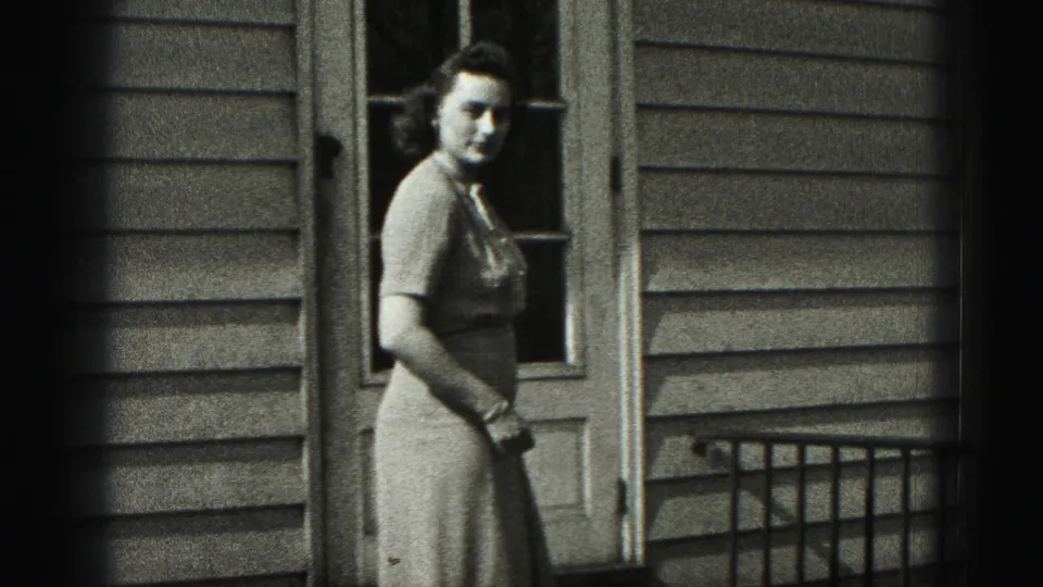 The woman stands beside the house door and looks toward the camera in black-and-white 8mm footage from the Harrisburg metadata.