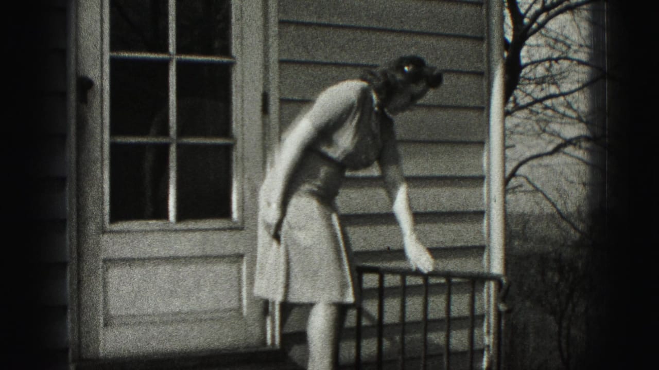 In 1975 Harrisburg home-movie footage, a woman in a dress bends near a porch railing beside a paneled door and house siding.