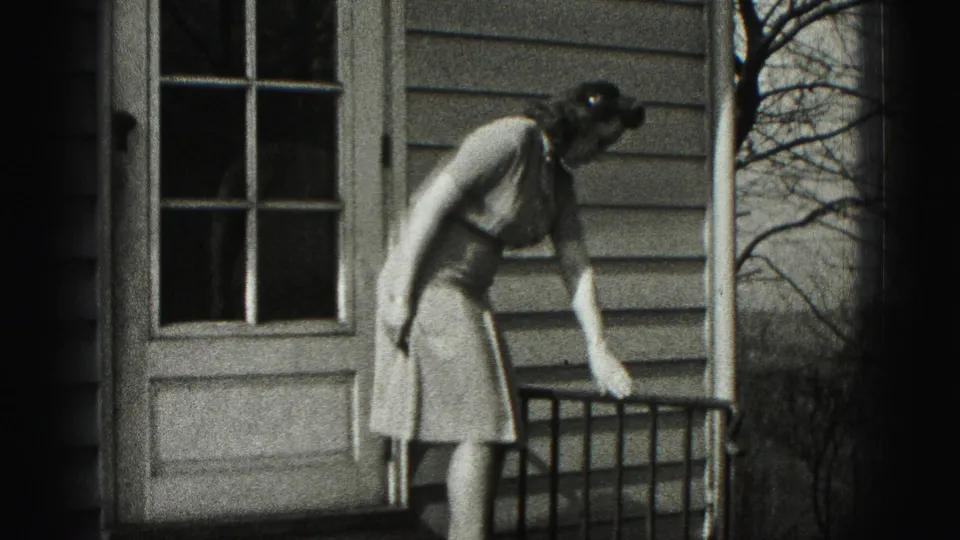 In 1975 Harrisburg home-movie footage, a woman in a dress bends near a porch railing beside a paneled door and house siding.