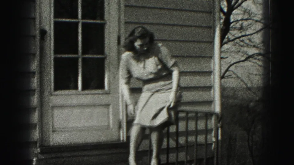 The woman sits on the porch railing near a glass-paneled door, with house siding and bare branches visible behind her.