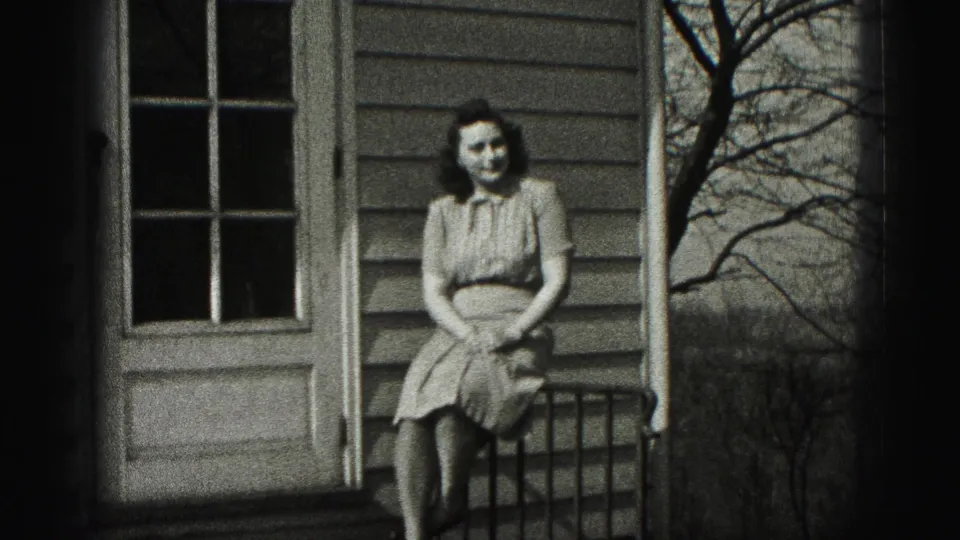 A woman in a light dress sits facing the camera on a porch railing outside a residential house in the Harrisburg clip.
