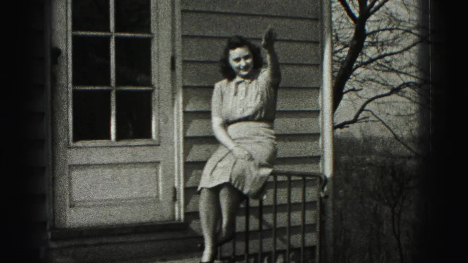 The woman smiles and waves while seated on the porch railing, framed by house siding, a door, and bare tree branches.