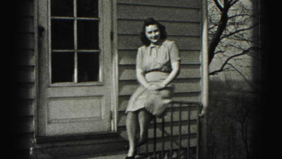 In Harrisburg, Pennsylvania, a woman sits on a porch railing in a light dress, smiling toward the camera in 1975 home-movie footage.