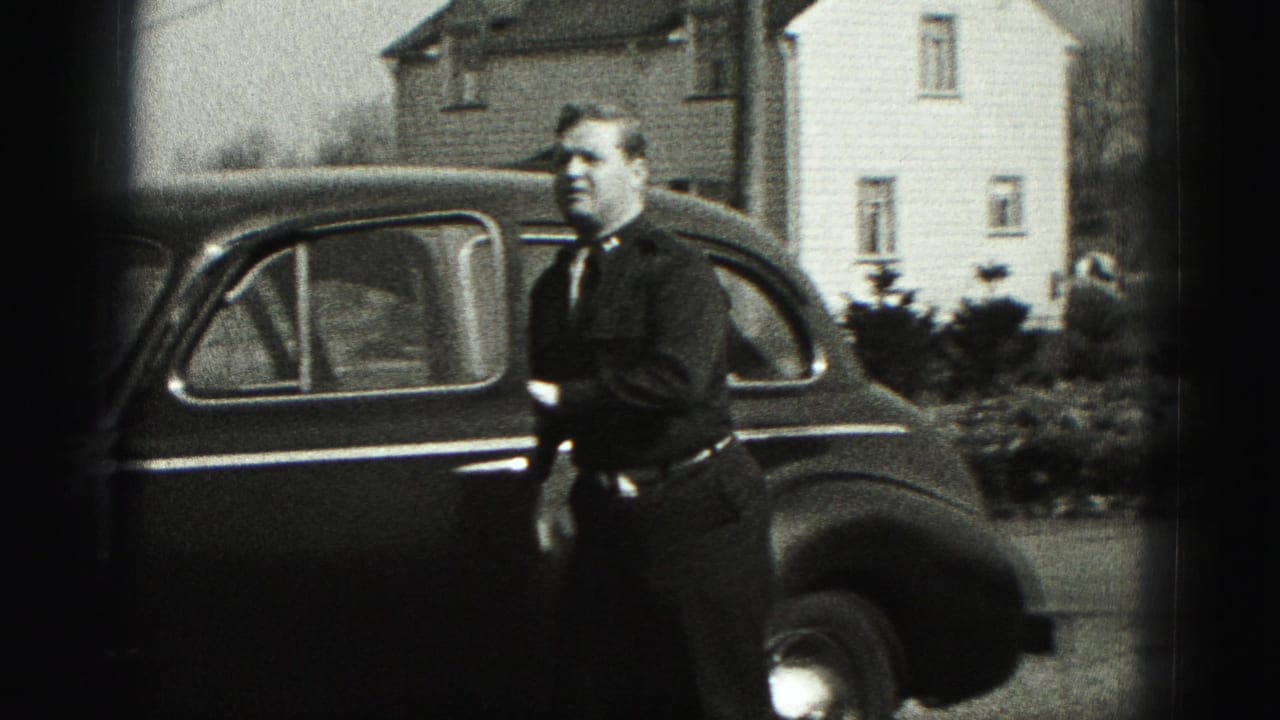 A man stands next to a dark mid-century sedan with houses and shrubs behind him in the Harrisburg home-movie scene.