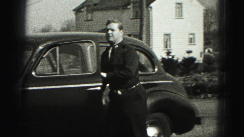A man stands next to a dark mid-century sedan with houses and shrubs behind him in the Harrisburg home-movie scene.