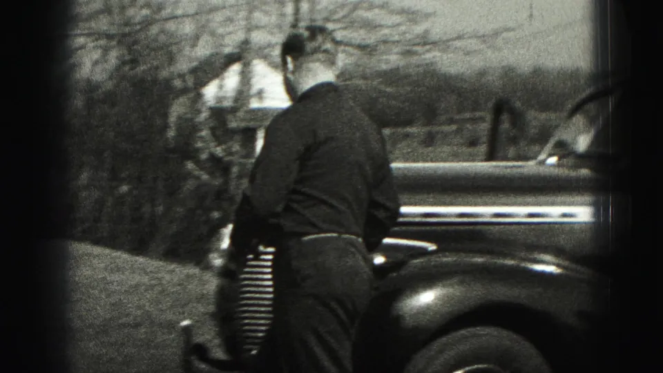 Seen from behind, the man stands near the sedan's front end while a house, lawn, shrubs, and bare trees appear in the background.