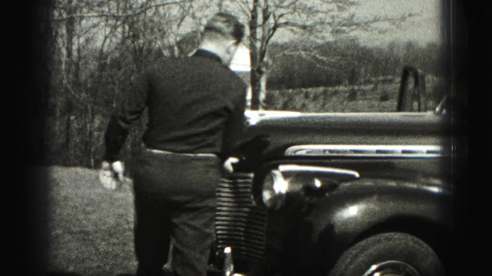 In Harrisburg, Pennsylvania home-movie footage from 1975, a man stands by the front fender of an older car near bare trees.