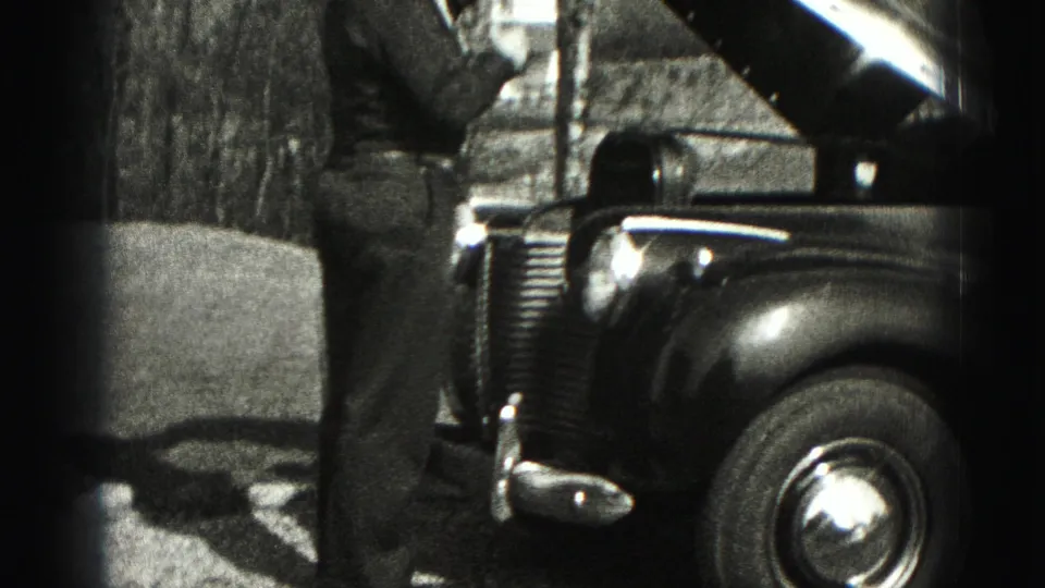 A man stands at the front of an older car with the hood lifted, filmed outdoors in 1975 Harrisburg-area home-movie footage.