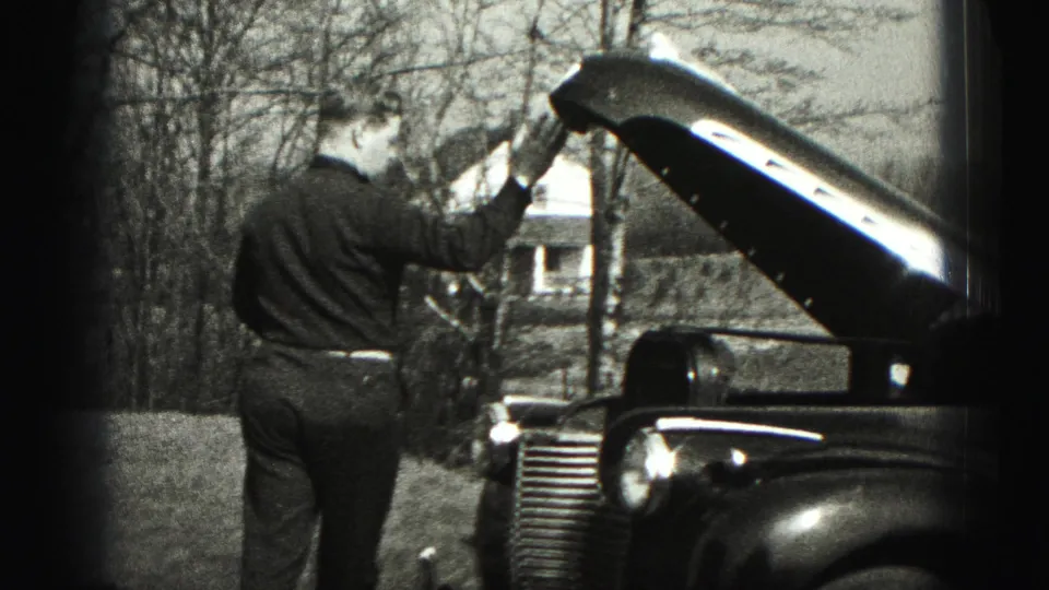 The man reaches up toward the open hood of an older car, with bare trees and a distant house behind him in Pennsylvania footage from 1975.