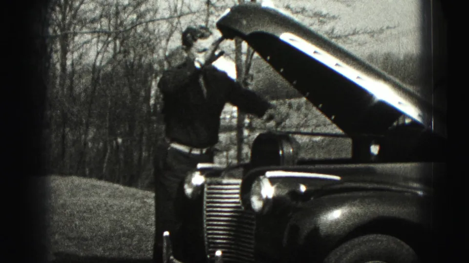 The man stands beside the raised hood and front grille of an older car in 1975 Harrisburg home-movie footage, with a rural background visible.