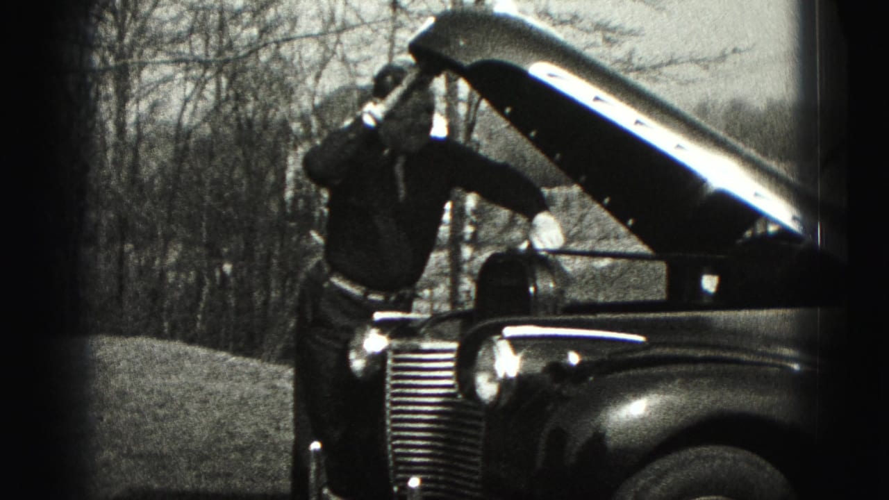 Man stands beside a vintage car with the hood raised in a wooded outdoor area.