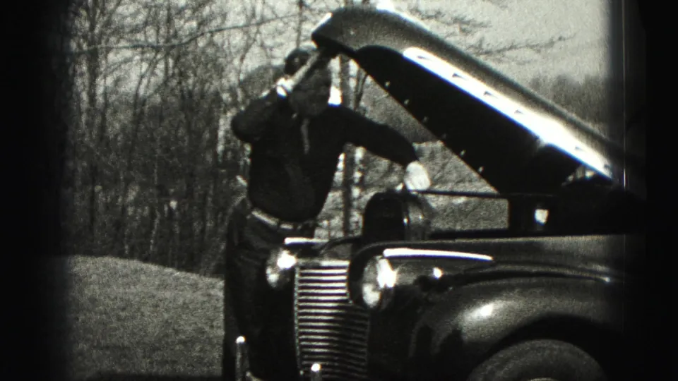In Harrisburg-area 1975 home-movie footage, a man works near the open hood of an older car with bare trees behind him.