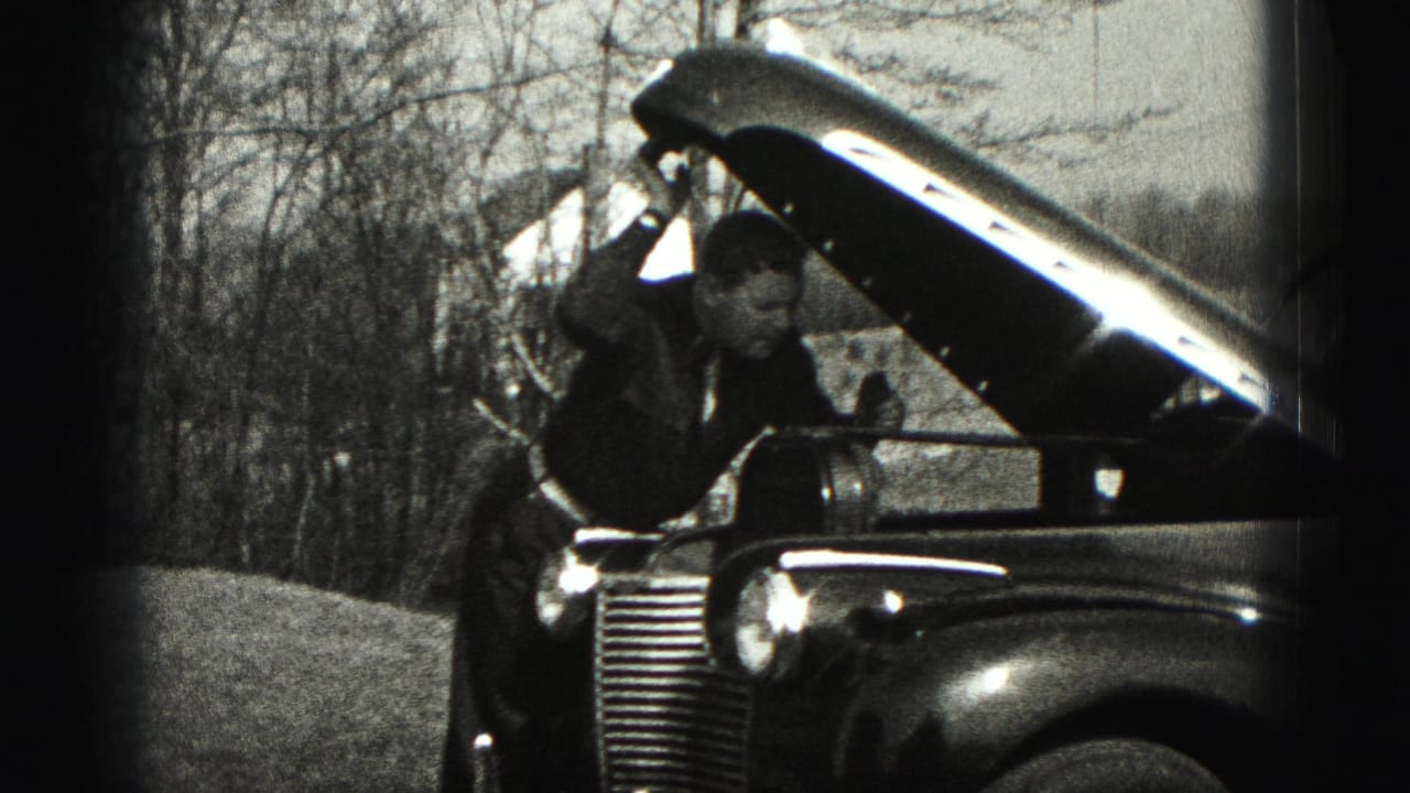 A man in dark clothing leans toward the engine compartment of an older car during a 1975 Harrisburg home-movie repair scene.