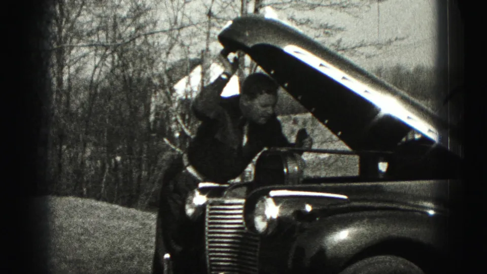 A man in dark clothing leans toward the engine compartment of an older car during a 1975 Harrisburg home-movie repair scene.
