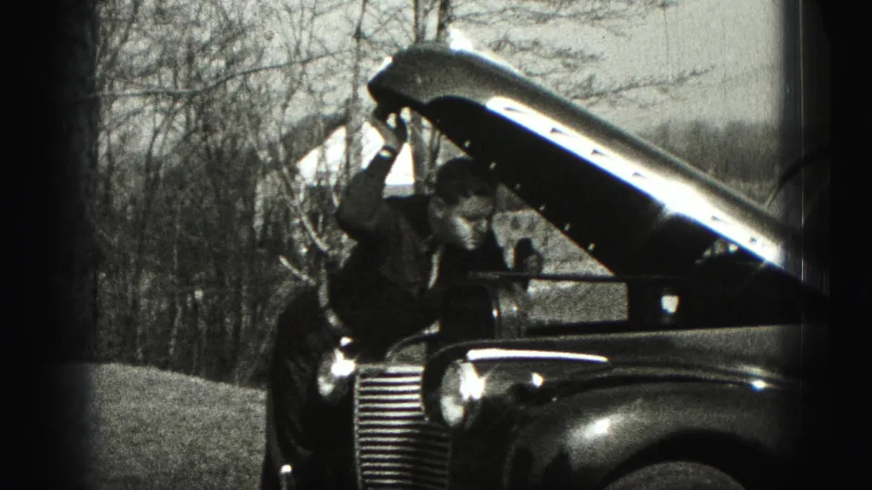 The man bends deeper into the open engine bay of a vintage car, with the rural Harrisburg setting visible behind him.