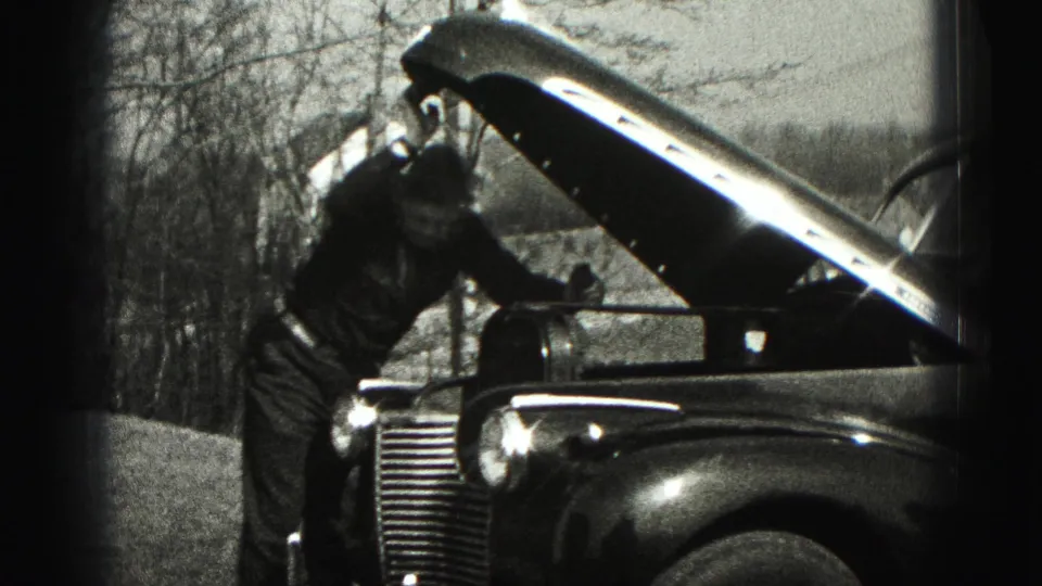 A 1975 home-movie frame shows a man reaching beneath the raised hood of an older car, likely continuing a roadside or driveway repair.